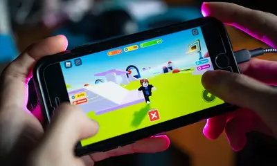 Hands holding a phone showing Roblox gameplay with a Roblox character on screen